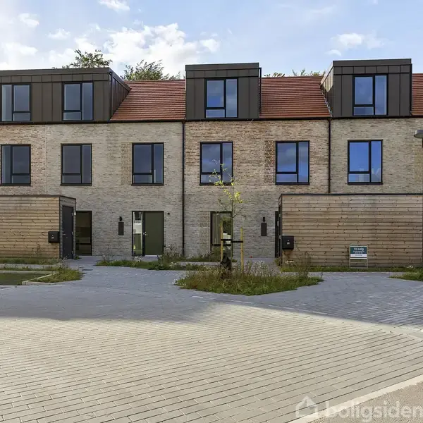 Rækkehuse med moderne arkitektur står på rad og række. Solstråler belyser de røde tegltage og store vinduer. En handicapparkeringsplads er markeret med et skilt foran husene.