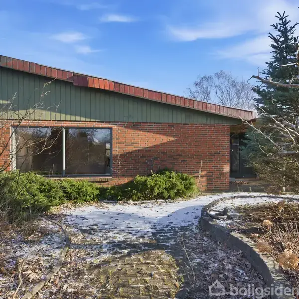 Et hus i mursten med store vinduer står omkranset af vinterbar buskads og et sneklædt haveanlæg. Huset har et skråt tag, og himlen er klar med få skyer.