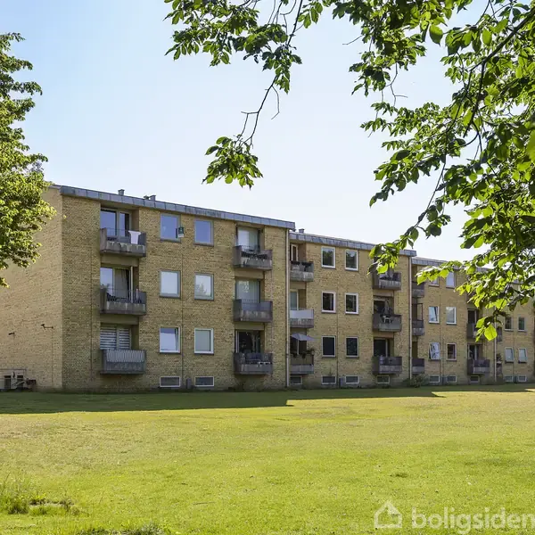 En række af gule murstensbygninger med altaner står stille i en grøn græsplæneomgivelse, omgivet af træer under en klar himmel.