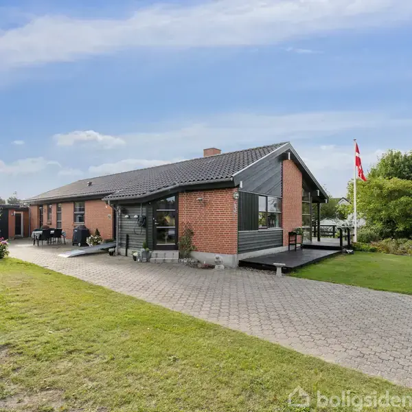 En murstensvilla med sort tag står i et grønt haveområde. Villaen har en stor terrasse og flagstang foran. Skyfri, blå himmel ses i baggrunden.
