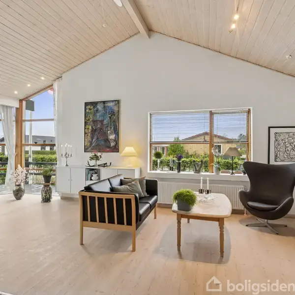 Stue med højt til loftet, sort lænestol, træsofa med hynder, og lille sofabord. Store vinduer giver udsigt til haven. Vægdekorationer og planter skaber en hyggelig atmosfære.