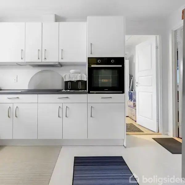 Hvidt køkken med skabe og indbygget ovn; en kaffemaskine står på bordet. Et køleskab med isdispenser er til højre. Væggene er lyse, og der er åbning til en gang.