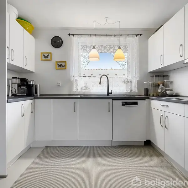 Køkken med hvide skabe står stille. På væggen hænger tre billeder og et ur. En vask med armatur står under et vindue med blondegardin. Bordpladen er mørk.