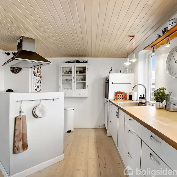 Et lyst køkken med træloft og -gulv indeholder en lang køkkenbordplade. Køleskab, komfur og vask er integreret. Vægur og lamper pryder væggen, mens en trappe ses i baggrunden.