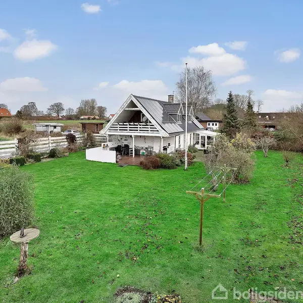 Et hvidt hus med skråtag står centralt i en stor grøn have. Omkring huset ses træer, buske og en græsplæne. I baggrunden er der marker og flere træer.
