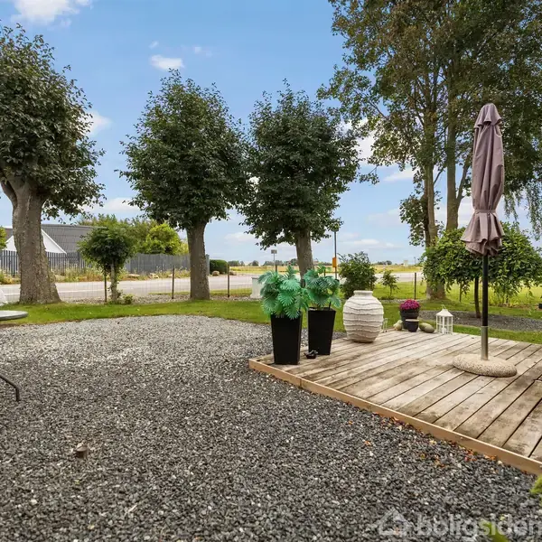 Grusbelagt terrasse med stole og borde, omkranset af træer og en lille græsplæne. En parasol står på en træplatform. Udsigt til en vej og åben mark i baggrunden.