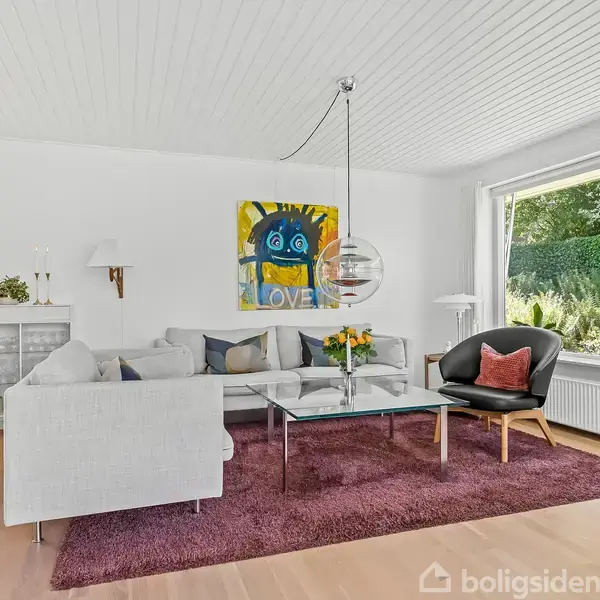 Stue med hvid sofa og sort lænestol ved et glasbord med blomster. Kunstmaleri på væggen. Store vinduer giver udsigt til grøn have. Tæppe på trægulvet.