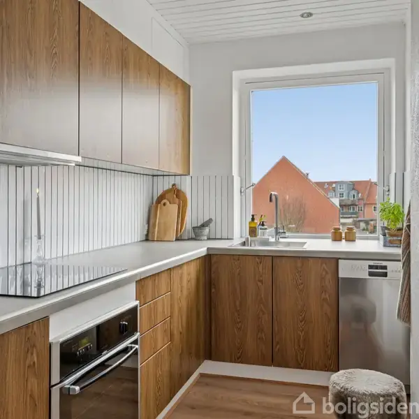 Et moderne køkken med træskabe og en hvid bordplade. En indbygget ovn og kogeplade. Vindue med udsigt til røde hustage og blå himmel.