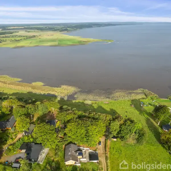 Flod breder sig ud i landskab omgivet af grønne marker og små huse. Området viser en landlig idyl med skovområder tæt ved vandkanten, hvor både stille ligger ved en mole.