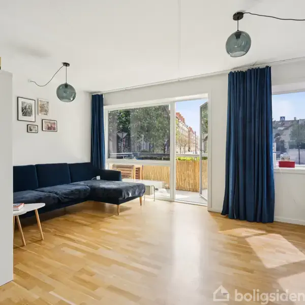 En blå sofa står i en stue med lyse trægulve. Rummet har store vinduer og blå gardiner, der giver udsigt til en balkon og træer udenfor.