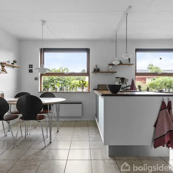 Et moderne køkken med spisekrog. Køkkenbordet har indbyggede skabe og hængende lamper over. Spisebord med sorte stole står ved væggen. Vinduet afslører en frodig, grøn have udenfor.