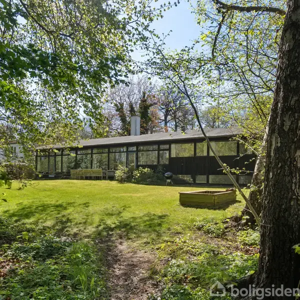 Et moderne hus med store vinduer står i en frodig have. Træer og buske omgiver græsplænen og skaber en naturskøn atmosfære i et skovområde.