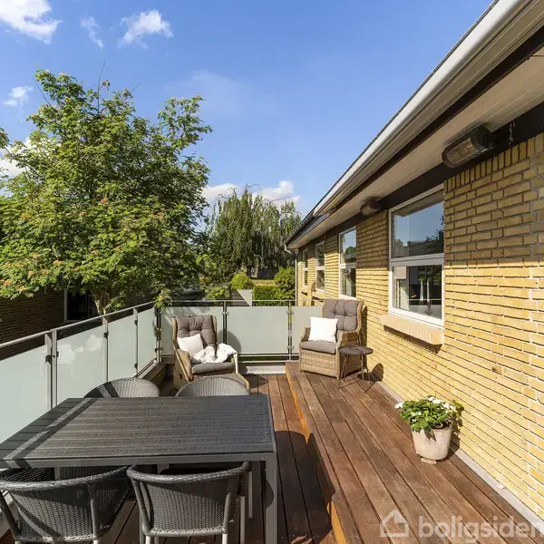 Terrassens hovedfokus er et sort havebord med stole, omgivet af en gul murstensvæg og glasrækværk, beliggende ved et træ i en solrig have.