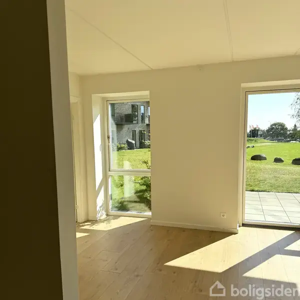 Rum med vinduer til græsområde, der tillader dagslys at strømme ind, hvor et større vindue står åbent mod terrassefliser. Udenfor ses en boligbygning og sten på græsset.