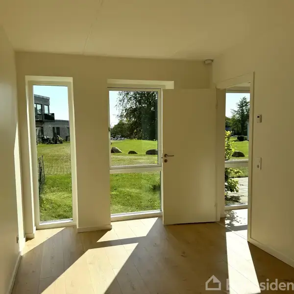 En åben dør med glasruder lukker sollys ind, og afslører en græsklædt have med buske og træer udenfor. Indendørs er der lyse vægge og trægulv.