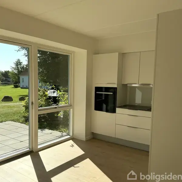 Køkken med hvide skabe og indbygget ovn står stille i et lyst rum. En stor glasdør åbner ud mod en grøn have med træer og terrassesten.