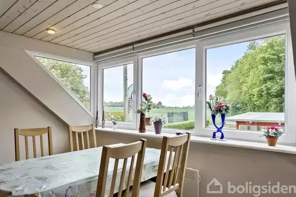 Et spisebord med træstole står ved store vinduer. Vaser med blomster pynter vindueskarmen, mens udsigten viser frodige træer og marker i et lyst rum.