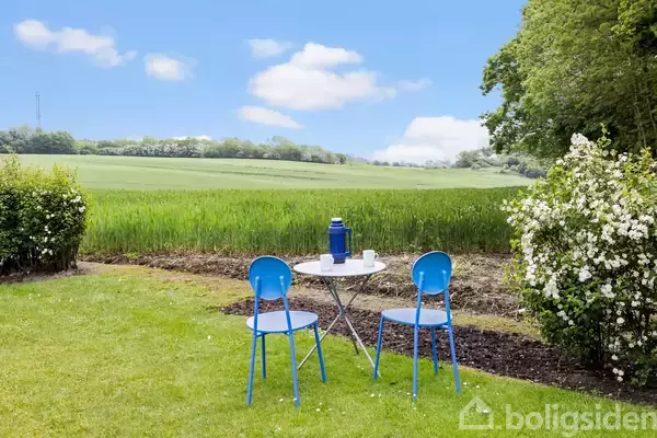 To blå stole og et rundt bord med en blå termokande står på en græsplæne. Omgivelserne er en grøn mark og frodige buske under en blå himmel.