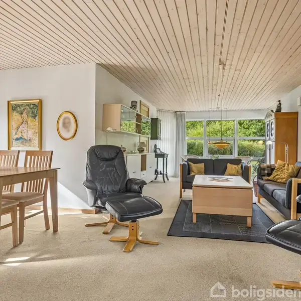 Stue med sort sofa, lænestole og træbord placeres foran store vinduer. Spisebord med stole til venstre. Træloft og kunstværker pryder væggene, lys strømmer ind.