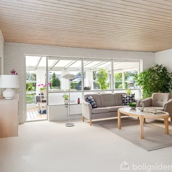 En stue med sofaer og et sofabord står midt i rummet. Store vinduer giver lys og udsigt til en terrasse med planter udenfor.