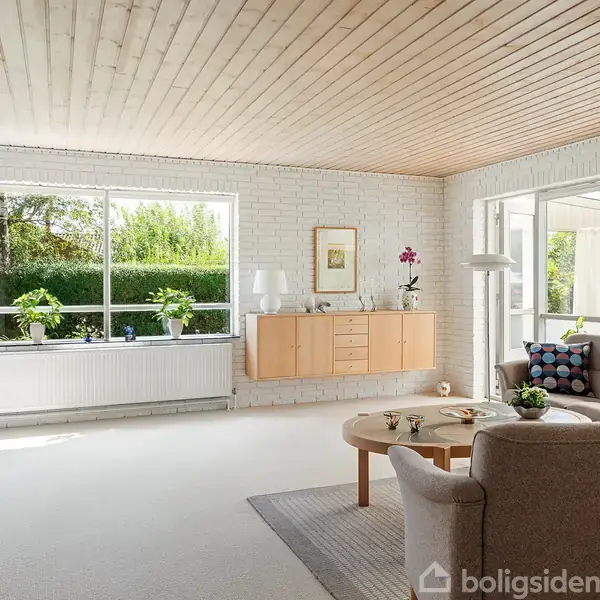 Stue med lyse møbler; to sofaer og et rundt bord står på et tæppe. Store vinduer lader lys ind og viser grønne haver udenfor. Væggene er hvide mursten.
