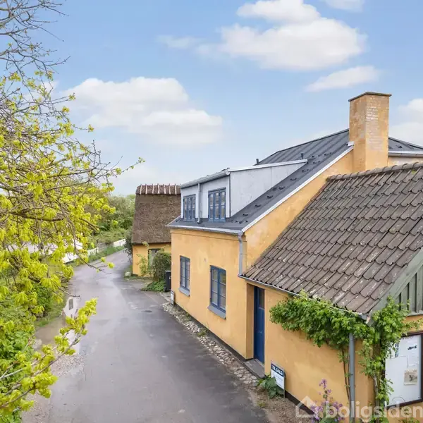 Gult hus med skråtag, omgivet af en stille vej og grønne træer under en let overskyet himmel. Huset har blå vinduer, og der vokser klatreplanter ved indgangen.