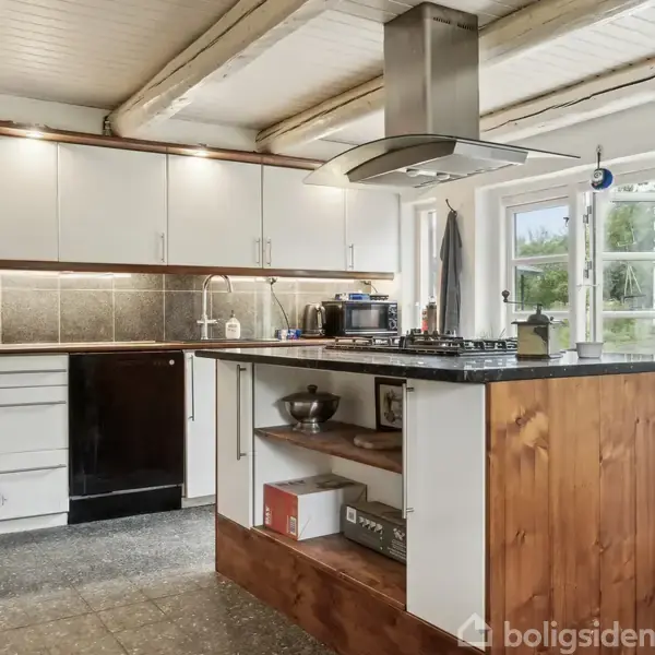 Et køkken med en central ø, hvor en kogeplade og emhætte er placeret. Vægskabe og skuffer omkranser rummet, og store vinduer giver udsigt til haven.