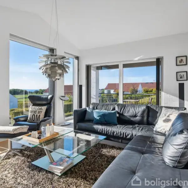 Stue med sort lædersofa og lænestol omkring et glas-sofabord, belyst af naturlig sollys fra store vinduer, der viser udsigt til nabolaget. En moderne lampe hænger fra loftet.