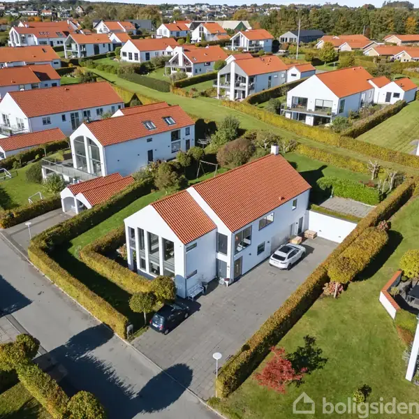 Hvide, moderne huse med røde tage ligger i et velordnet boligkvarter. Biler er parkeret på en brolagt indkørsel, omgivet af trimmede hække og velplejede haver under en klar, blå himmel.