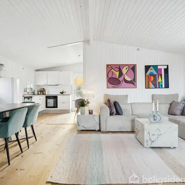 Sofagruppe med grå sofa og marmorbord, dekoreret med puder, placeret i en lys stue. Til venstre står spisebordet med blå stole. Væggene har farverige kunstværker.