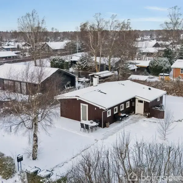 Et sneklædt hus med brune vægge står i et snedækket landskab med flere omkringliggende boliger og træer. Formidler en rolig vinterstemning.