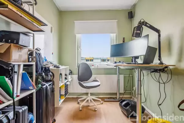 Stol foran et hæve-sænke-bord med computer og skærm i et kontor. Omgivet af reoler fyldt med bøger og papirer. Vinduet i baggrunden slipper lys ind.