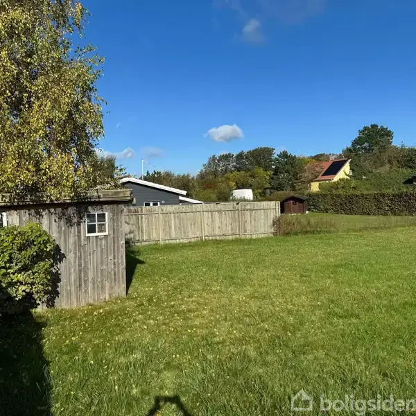 En træhytte står stille i en grøn have omgivet af et hegn. Baggrunden er fyldt med huse under en klar blå himmel med spredte skyer.