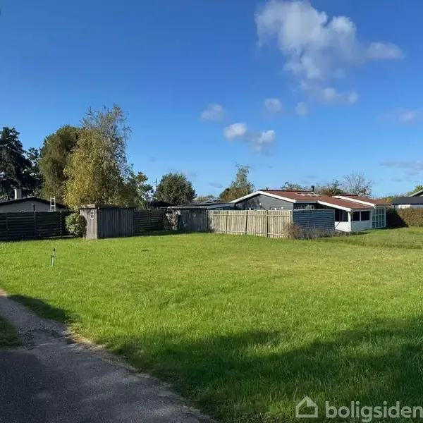 Grønt græsareal strækker sig foran en hvid bygning med rødt tag. Bygningen er omgivet af et hegn i et landligt miljø, under en klar blå himmel med få skyer.