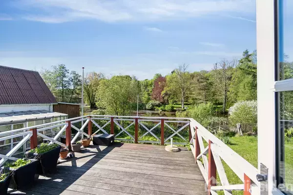 Terrasse med blomsterkrukker står stille foran en malerisk udsigt over en grøn have og en sø, omgivet af træer under en klar blå himmel.