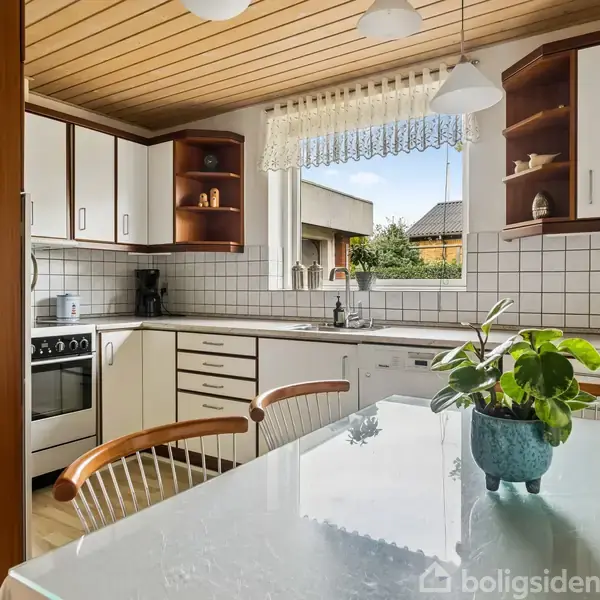 Køkken med L-formet bordplade, hvidt fliseskørt. Ved bordet står stole, og en plante er placeret på bordet. Lys træloft. Udendørs udsigt gennem vindue med blondegardin.