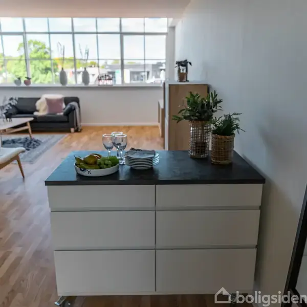 En kommode står i en stue. Ovenpå er glas, skåle, og planter. Stuen har parketgulve, en sofa, og store vinduer, der lukker naturligt lys ind.