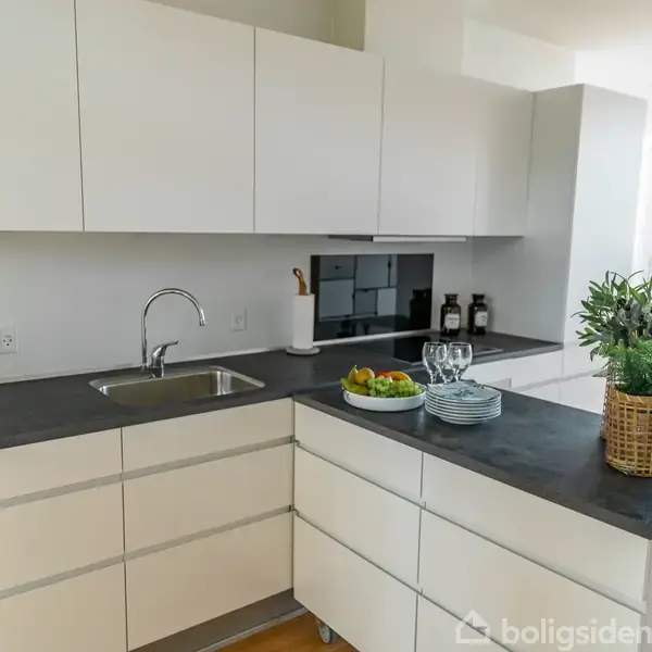 Et moderne køkken med hvide skabe og en sort bordplade. Der står planter, en skål med frugt og glas på bordet, mens en stue er synlig i baggrunden.