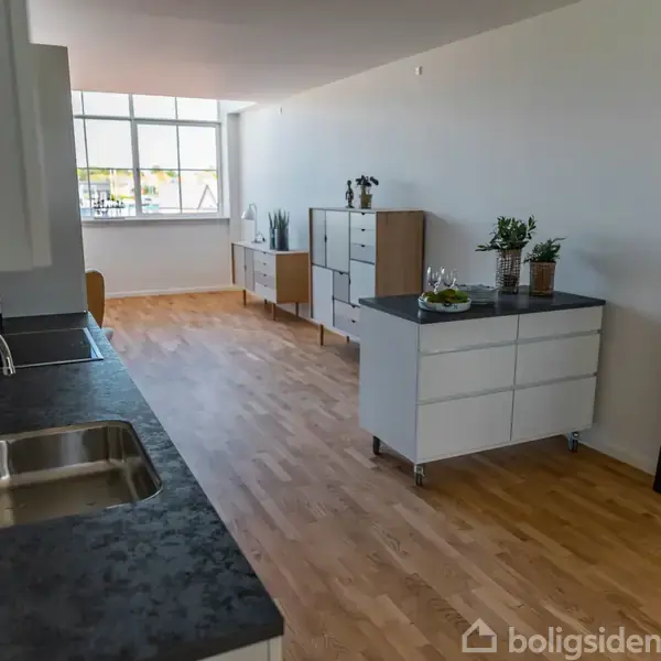 Køkken med sort bordplade og dobbeltvask. Rum med trægulv indeholder køkkenskabe, planter, stort vindue og spejl. Fra "Boligsiden".