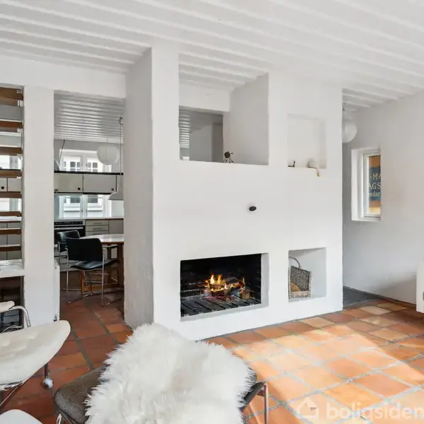En stue med en brændende pejs placeret i en hvid skillevæg. Omkring står moderne møbler, herunder en lænestol og en sofa. Trægulv og trapper ses i baggrunden.