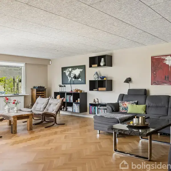 Stue med sort hjørnesofa og sofabord på trægulv. Store vinduer giver udsigt til haven. Der hænger kunst på væggen, og hylder med bøger og dekorationer er til stede.