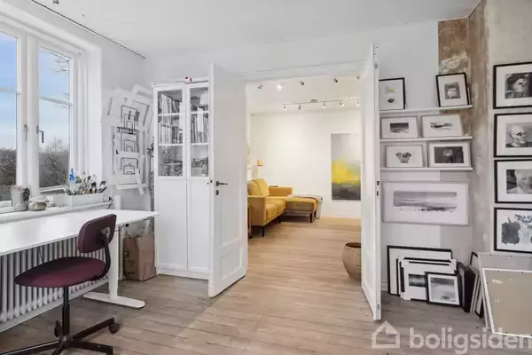 Et skrivebord med stol står ved et vindue med kunstmaterialer. Dørene fører til en stue med orange sofa. Væggene er dekoreret med billeder og tegninger.