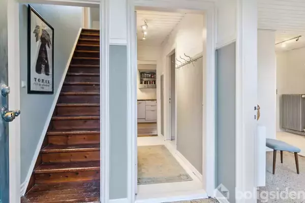 Trætrappe fører opad i en lys gang med hvidmalede vægge og grå paneler. Til venstre hænger en plakat med teksten "TOBAK". I baggrunden ses et køkken.