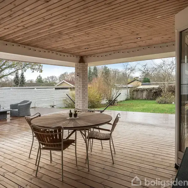 Rundt bord med stole står under overdækket terrasse, mens grå sofaer er placeret mod hegnet. Baggrunden viser en have med græsplæne og buske.