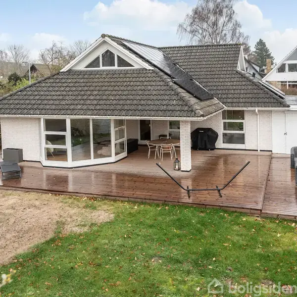 Et moderne hus står stille på en bred træterrasse. Haven er grøn og rummelig, med en hængekøje og kurvestole arrangeret under. Huset har store vinduer og muret tag.