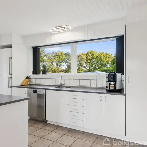 Køkken med hvide skabe og skuffer, sorte bordplader, kaffemaskine ved siden af vasken. Store vinduer viser udsigt af grønne træer. Flisefuld gulv skaber et lyst, luftigt miljø.