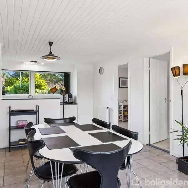 Et moderne spisebord med sorte stole står i et lyst rum. Rummet har hvidmalede trævægge, flisegulve og en sort reol. Et stort vindue giver udsigt til grønne omgivelser.