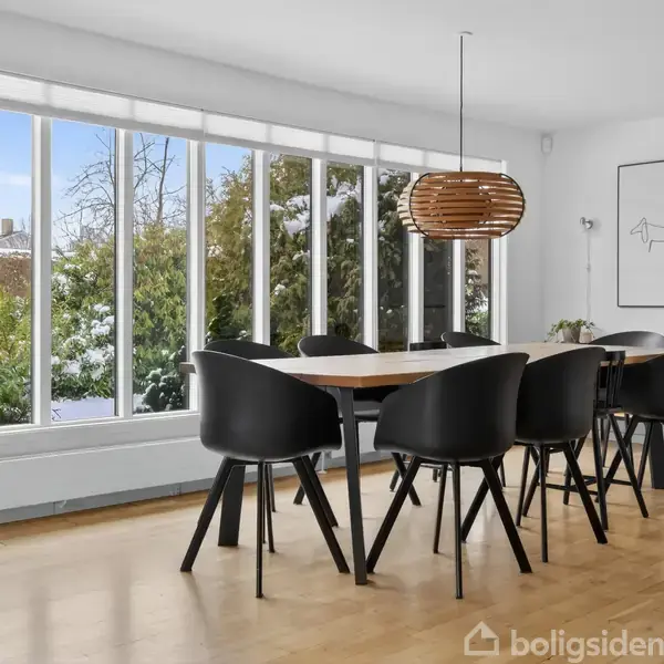 En spisebord med sorte stole står omgivet af store vinduer, der viser sneklædte træer udenfor. En moderne lampe hænger over bordet, og et minimalistisk billede pryder væggen.