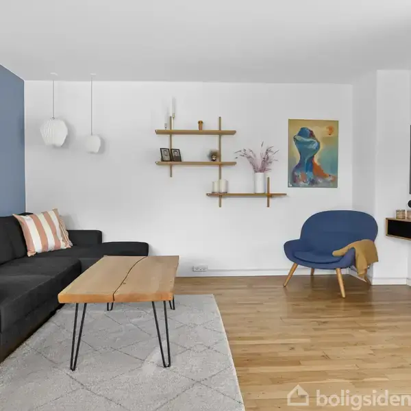 Sort sofa med stribede puder står ved en blå væg. Et træbord står foran. Blå stol i hjørnet ved stort TV. Hylder og kunst pryder rummet. Ingen tekst fundet.