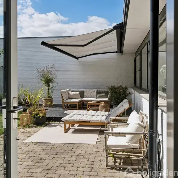 En terrasse med havemøbler, inklusive sofaer og stole, indrettet med puder og planter. En markise er opsat for skygge. Udsigt til grønt område gennem en åben glasdør.